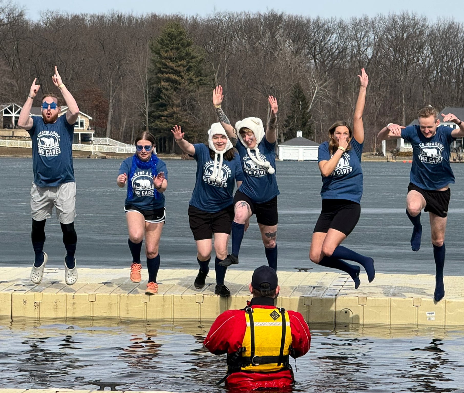 Six people in matching blue shirts, some wearing hats, jump enthusiastically off a dock toward a cold lake, as a person in a yellow and red safety vest stands in the water taking their photo. Trees and lakeside buildings are in the background.