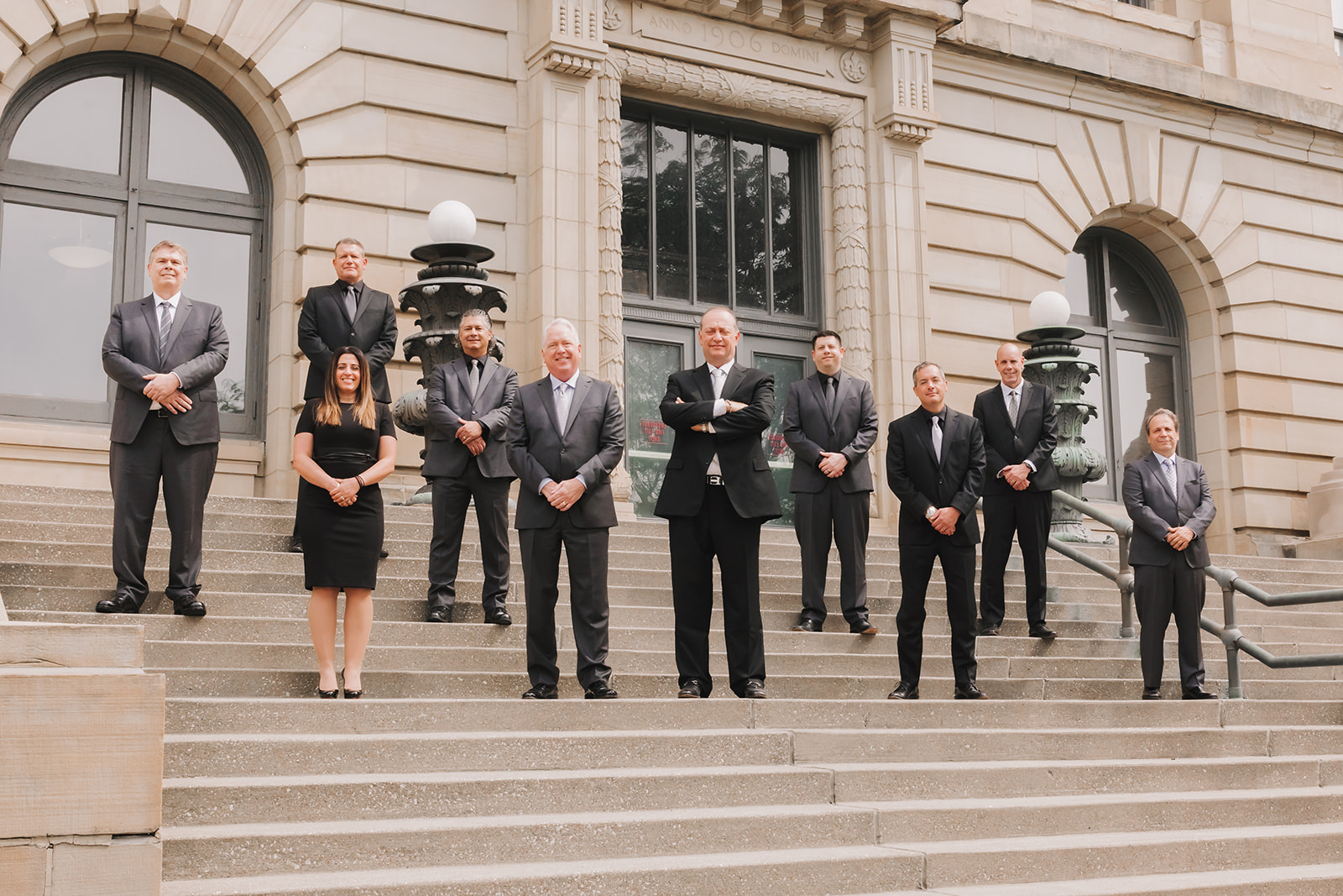 Nine people dressed in formal business attire stand spaced apart on the steps outside a large stone building with arched windows and lamps. Eight men and one woman face the camera with neutral expressions.