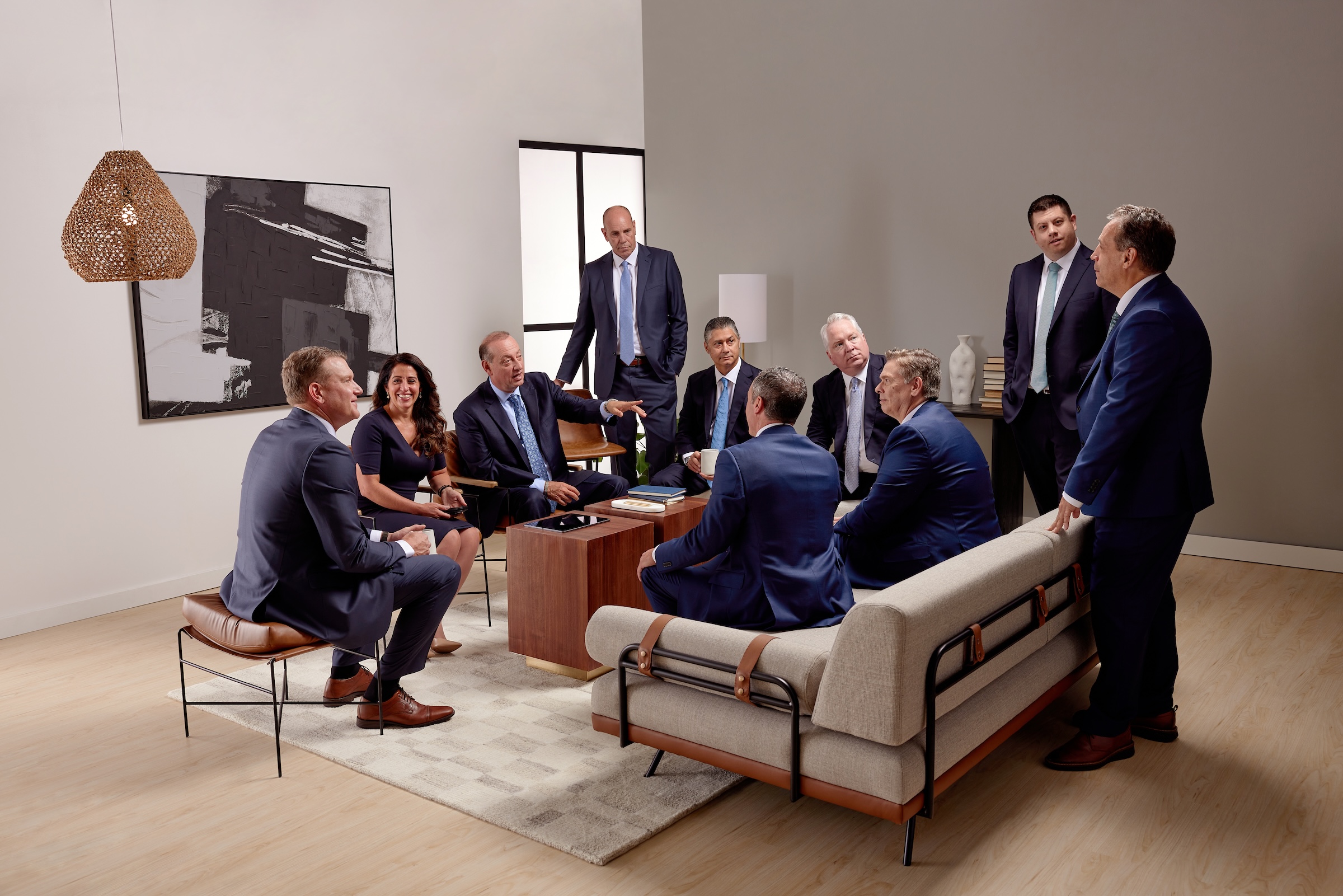 A group of nine people in business attire sits and stands around a coffee table in a modern office, engaged in discussion. The room features neutral colors, contemporary furniture, and a piece of abstract art on the wall.