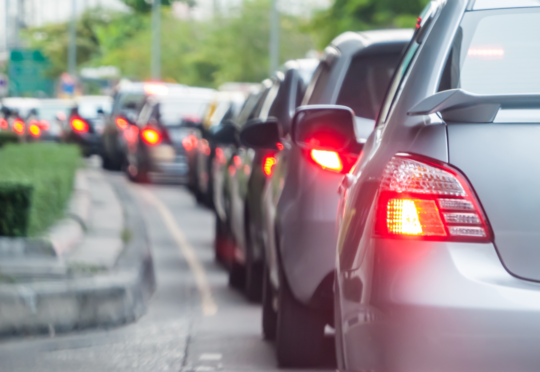 Una larga fila de coches está atascada en un atasco en una calle de la ciudad, con las luces de freno encendidas y la vegetación visible a lo largo del arcén.