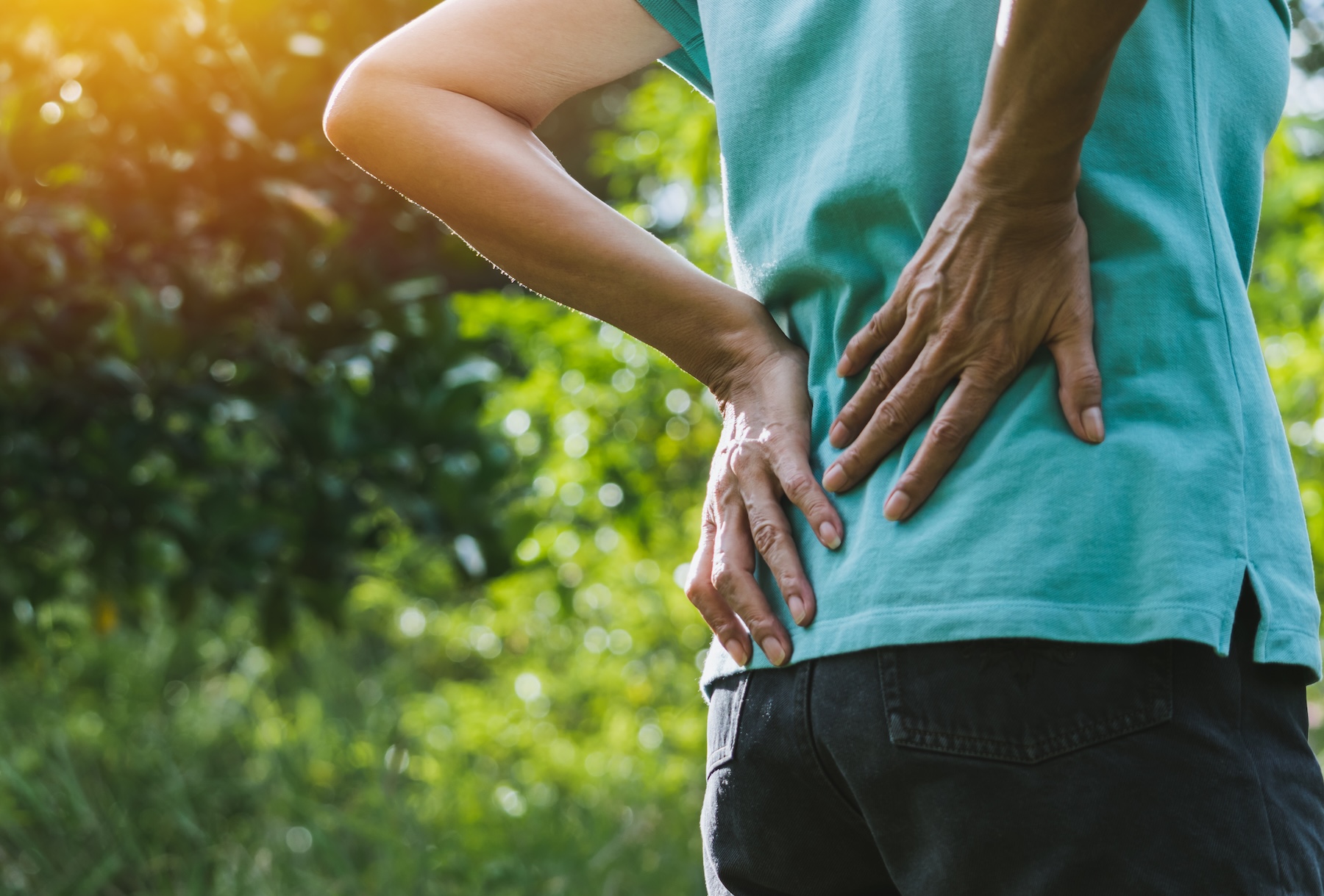 A person outdoors in a light blue shirt and dark pants is holding their lower back with both hands, appearing to experience back pain, with greenery and sunlight in the background.