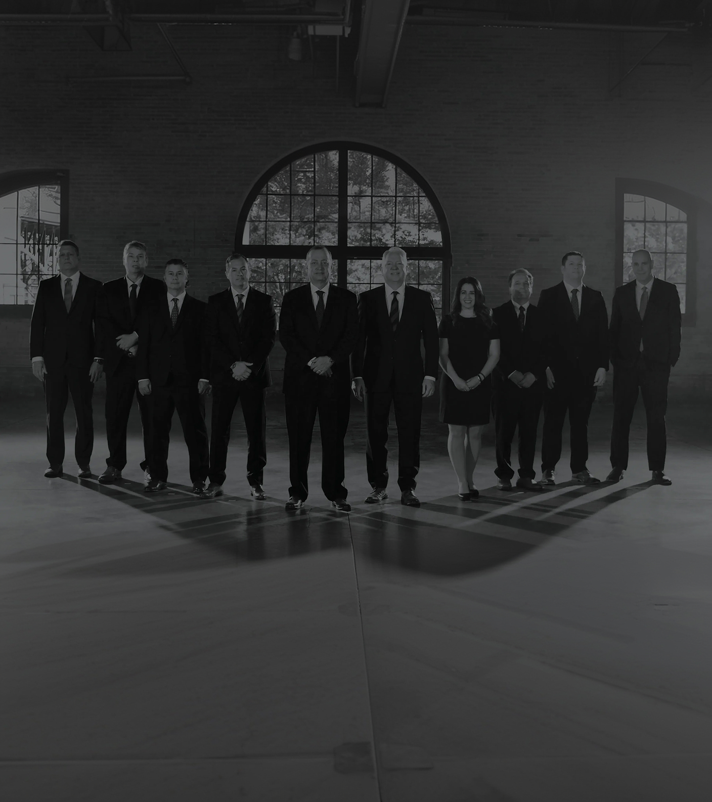 A group of nine men and one woman dressed in formal black attire stand in a spacious, industrial-style room with large arched windows and minimal lighting.