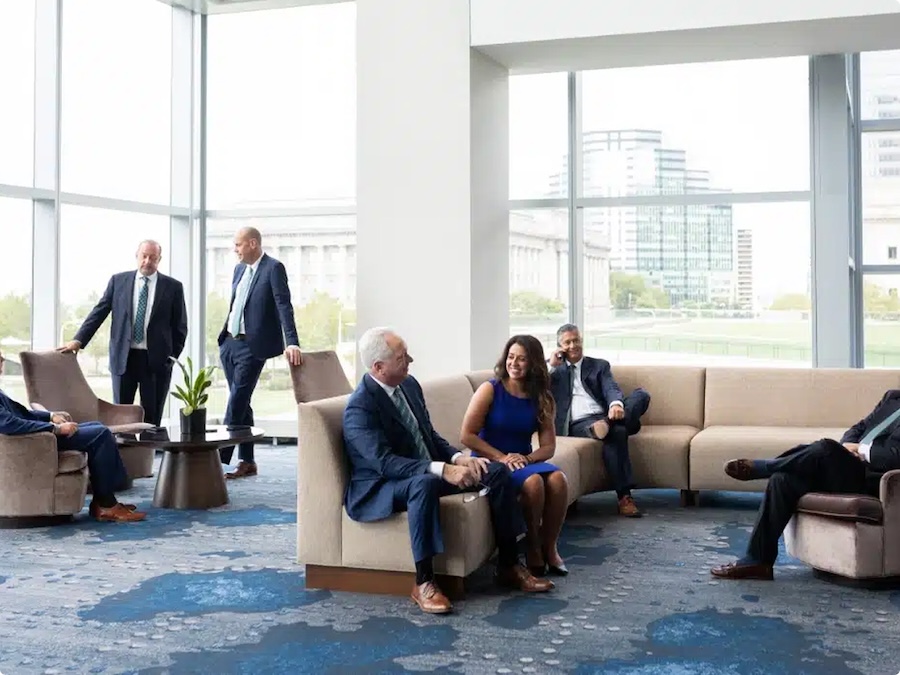 A group of business professionals, both men and a woman, sit and stand in a modern office lounge with large windows, natural light, and a cityscape view in the background. They appear to be engaged in casual conversation.
