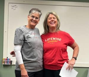 Two smiling women stand together in front of a whiteboard. One wears a gray “Kindness” shirt and glasses; the other wears a red “Lifewise Academy” shirt and holds a piece of paper. Colorful markers sit on the marker tray.