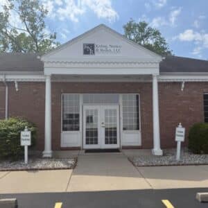 Brick office building with white columns and trim, featuring a glass double door entrance. Two signs in front read Visitor Parking. The sign above the door says Kisling, Nestico & Redick, LLC. Trees and blue sky in the background.