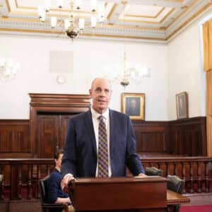 A man in a suit and striped tie stands at a wooden podium in a formal courtroom with chandeliers, wood paneling, and framed portraits on the walls.