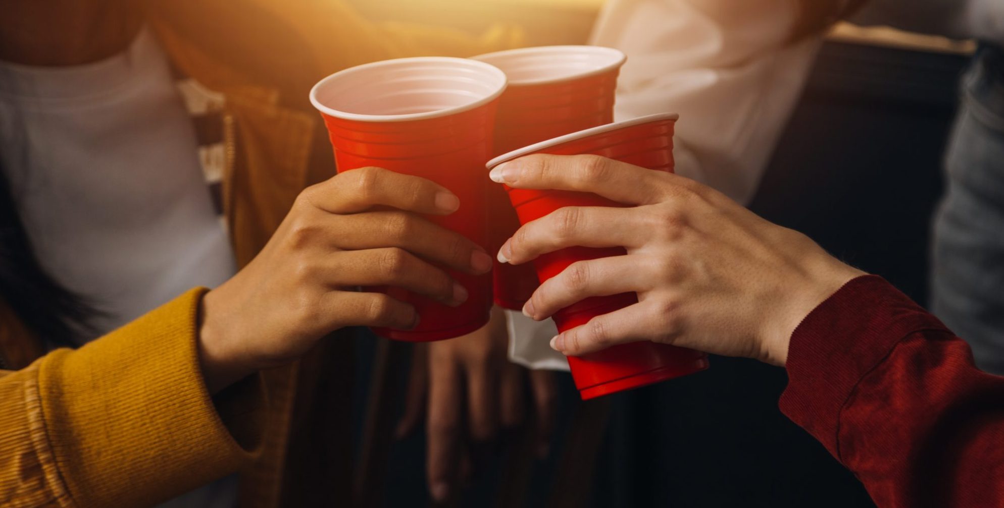 Three people clinking red plastic cups together in a toast, with warm lighting in the background. Only their hands and part of their arms are visible.