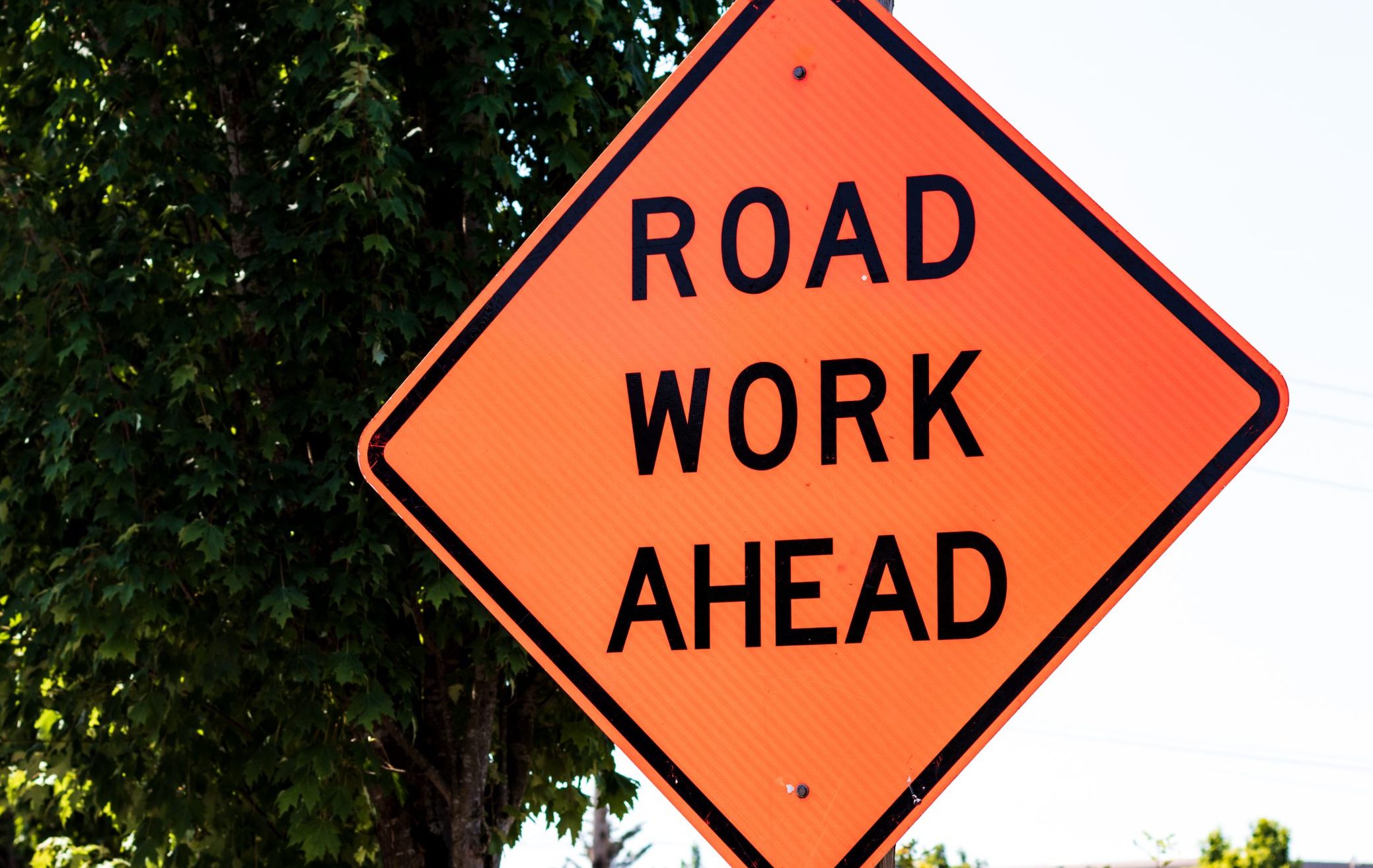 An orange diamond-shaped sign reads Road Work Ahead next to a tree-lined street on a sunny day.