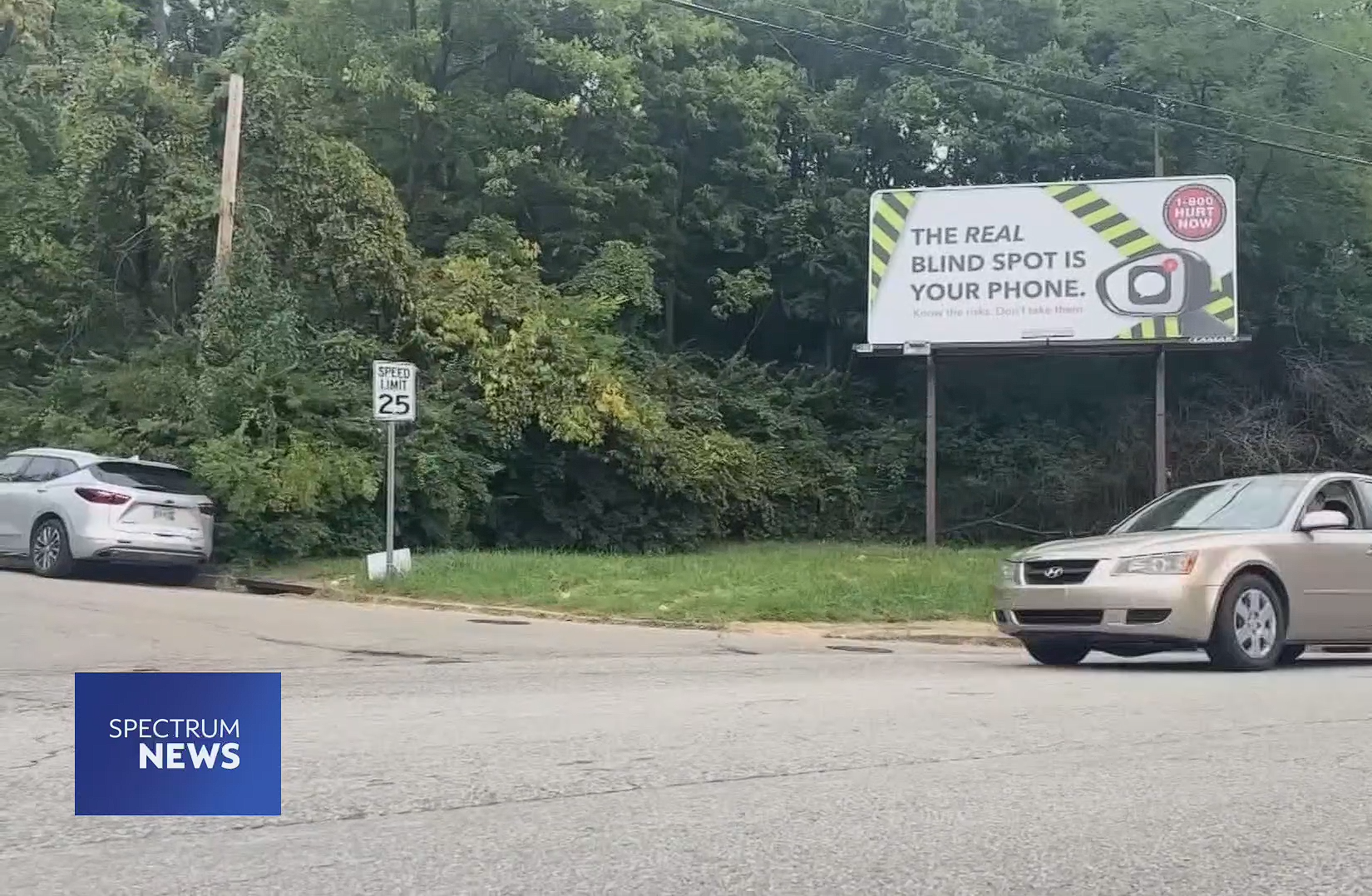 A car drives past a roadside billboard that reads, THE REAL BLIND SPOT IS YOUR PHONE, near a wooded area. A speed limit sign showing 25 mph and a Spectrum News logo are also visible.