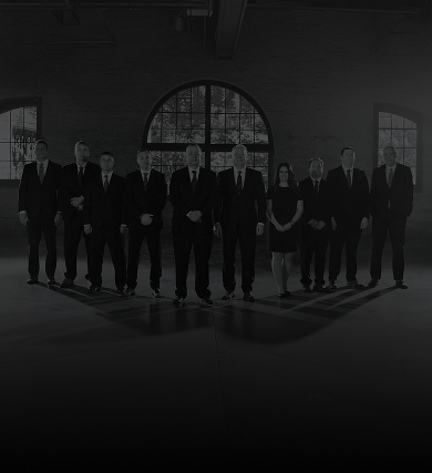 Ten people, mostly men in suits and one woman in a dress, stand in a dimly lit industrial room with large windows, casting long shadows on the floor.