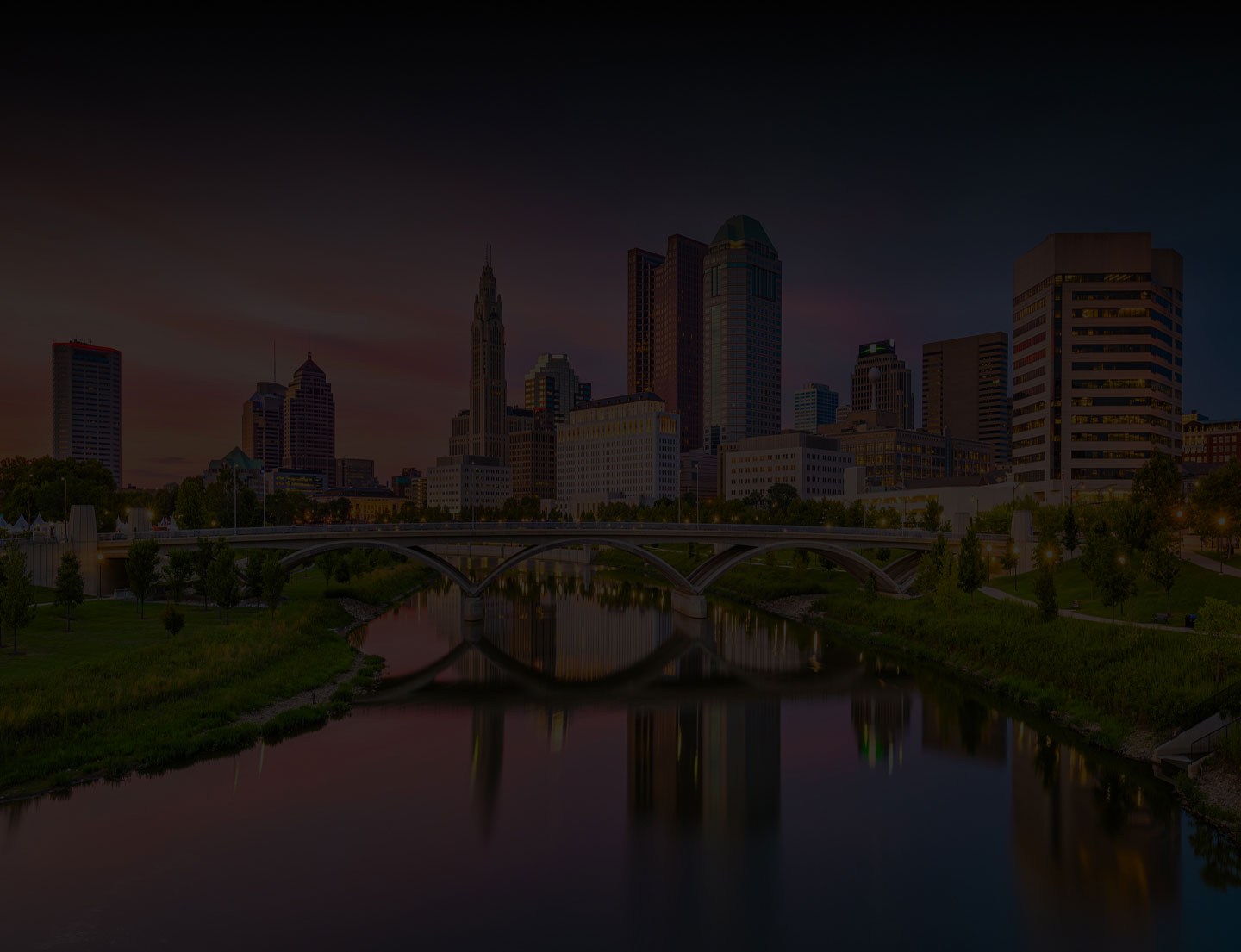City skyline at dusk with tall buildings and a bridge reflected in a calm river, surrounded by green trees and grass under a colorful evening sky.