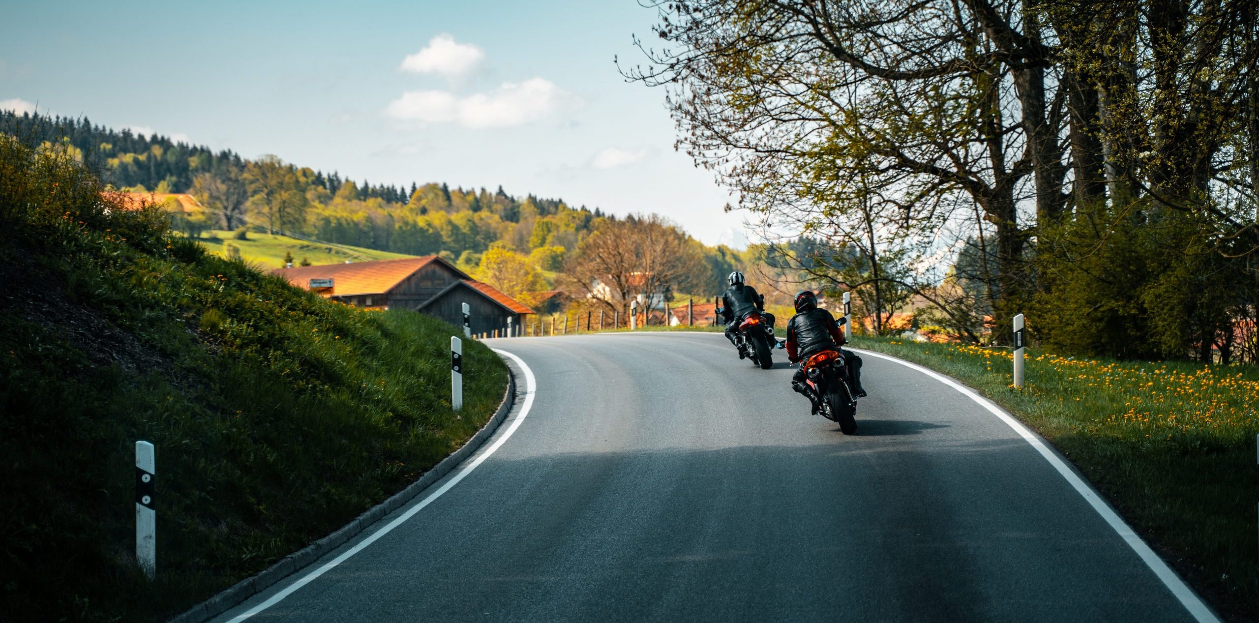 Two motorcyclists ride along a winding country road past grassy hills and trees.